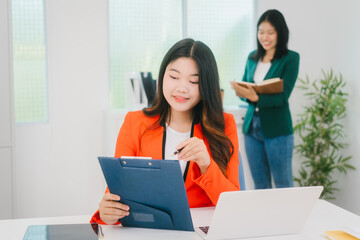 Two Asian business woman are discussing business reports and analyzing data at a desk. One is using...
