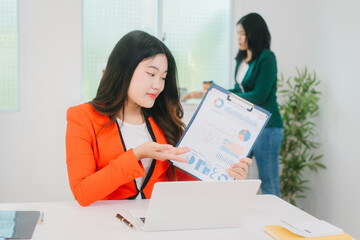 Two Asian business woman are discussing business reports and analyzing data at a desk. One is using...
