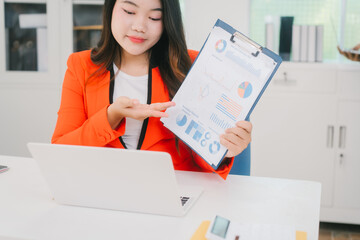 Two Asian business woman are discussing business reports and analyzing data at a desk. One is using...