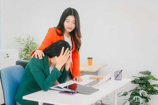 A stressed businesswoman holds her head in frustration while her colleague offers support by placing a comforting hand on her shoulder. They are working in an office with laptops and tablets