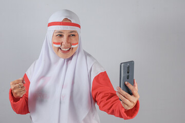 Muslim woman in red and white outfit smiling while holding smartphone and making a victory gesture, celebrating Indonesian Independence Day with joyful energy and digital engagement