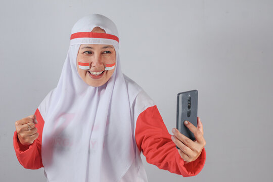 Muslim woman in red and white outfit smiling while holding smartphone and making a victory gesture, celebrating Indonesian Independence Day with joyful energy and digital engagement