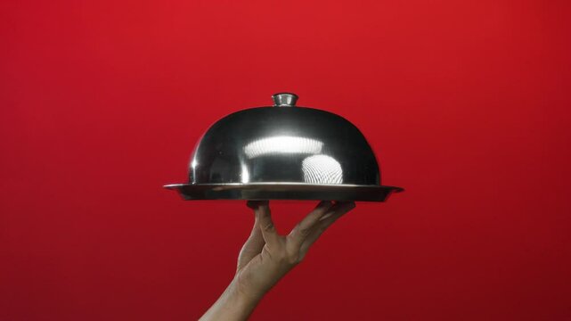 Hand holding a shiny cloche against a vibrant red background, showcasing a metallic serving presentation in an isolated minimalist setting.