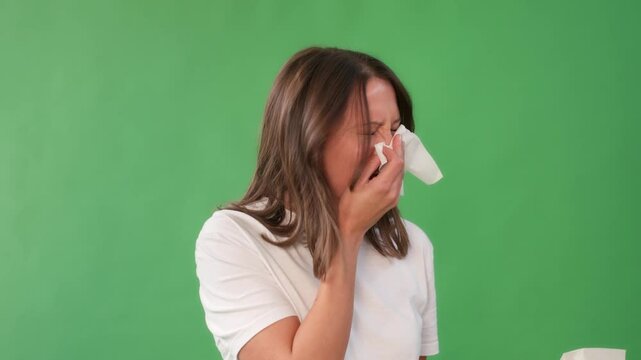 A woman with long hair  sneezing and wiping her nose with a napkin against a green background.