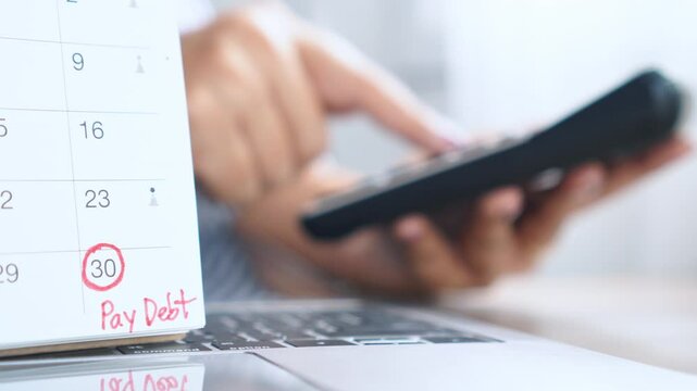 Close-up of a calendar with a reminder note to pay debt, with a woman's hand using a calculator in the background, showing personal finance planning and monthly bill tracking at home
