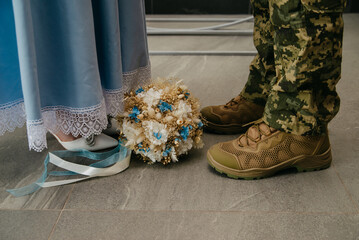 A wedding bouquet of colorful flowers with decorative inserts in it lies between the legs of the bride in a blue dress and the groom dressed in a Ukrainian military uniform