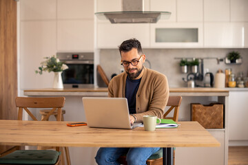 Obraz premium Bearded Man In Tan Cardigan Using Computer At Wooden Table In Modern Kitchen Interior