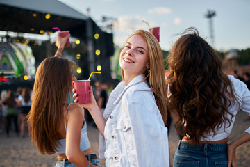 Women enjoy drinks at beach music fest, friends gather for fun, dance, summer vibes. Sunset party atmosphere, celebrate festival season with cold beverages, sea backdrop.