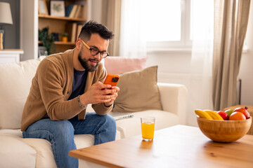 Smiling Man with Glasses Using Phone in Cozy Living Room with Warm Decor and Natural Lighting