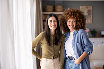 Two Diverse Young Women Standing Together In Naturally Lit Contemporary Living Room With Casual...