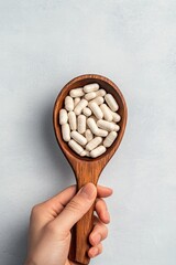 Close-up of a hand holding a wooden spoon full of white supplement capsules on a light grey background, symbolizing health and wellness.