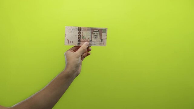 Man holding a saudi arabian riyal banknote against a bright yellow background, showcasing currency details with clear focus on the hand.