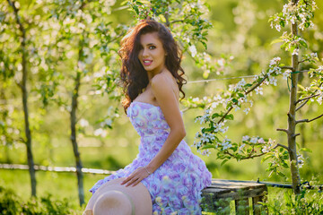 portrait of a beautiful woman in a dress sitting on a bench in a blooming apple orchard with a...