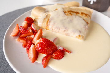 Side view of a plate with crepe pancakes, cut strawberries and vanilla sauce on grey coaster with a coffee cup in the back