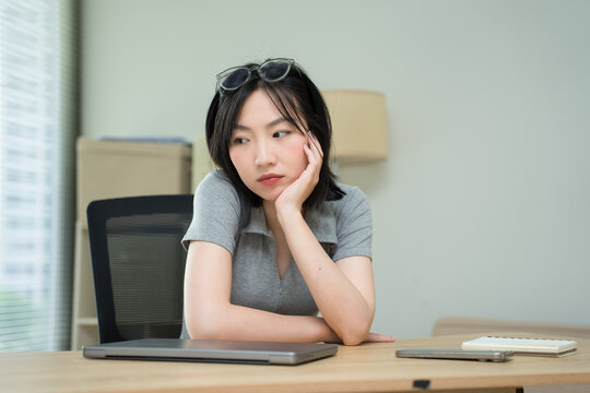 Young Asian woman thoughtfully sitting at a desk, reflecting on her work and the challenges of remote learning during the day in a modern office space
