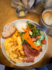 Tasty breakfast omelet with salad, bacon, curd and break on the side with cafe latte in the corner, top view