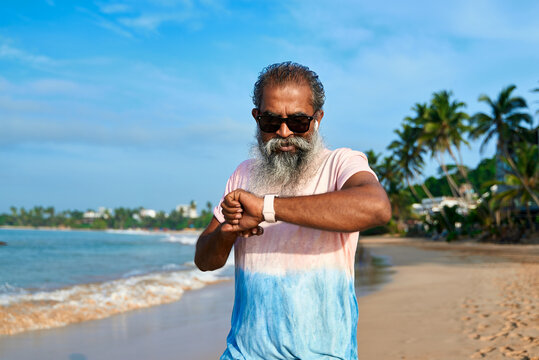 Senior ethnic man with beard uses smart watch on sunny beach. Modern tech tracks health stats. Wireless earbuds connected. Older generation enjoying gadgets. Tech-savvy grandpa into wearable devices.