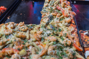 Close-up view of various delicious pizza slices with different toppings presented on trays behind a glass display counter. Highlights a tempting selection of prepared food ready to be served.