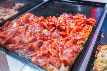Close-up view of various delicious pizza slices with different toppings presented on trays behind a glass display counter. Highlights a tempting selection of prepared food ready to be served.