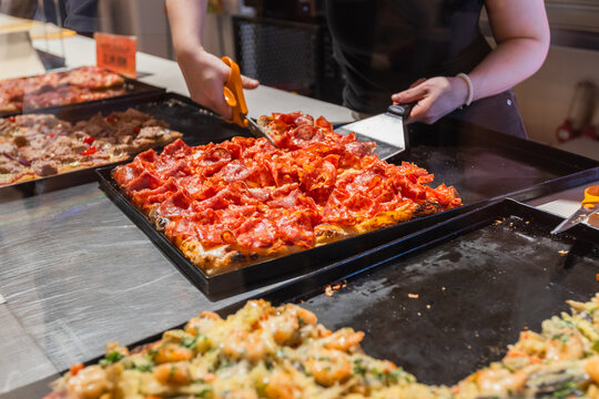 Close-up view of various delicious pizza slices with different toppings presented on trays behind a glass display counter. Highlights a tempting selection of prepared food ready to be served.