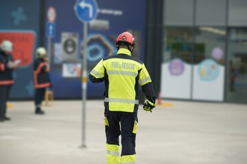 A fire and rescue worker in neon safety jacket and red helmet walks toward a glass fronted storefront while two colleagues in protective gear stand in the background on concrete pavement