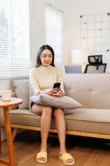 Fototapeta premium Beautiful Young Asian woman using phone on sofa at home, shopping online with internet in happy mood and Relax part time concept 