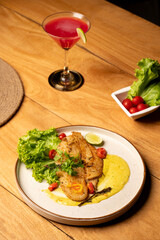 Pan-seared fish served on a plate with lettuce, cherry tomatoes, and sauce, placed on a wooden table with a red cocktail in the background for a refined seafood presentation.