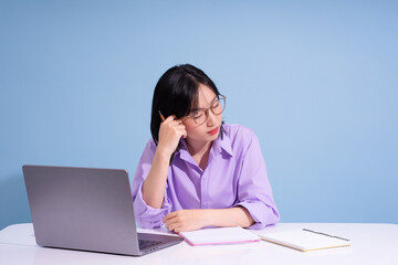 Young woman in a purple shirt sits at a desk with a laptop and notebooks, appearing deep in thought...