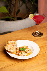 Prawns chimichurri served with bread on a plate, accompanied by a red cocktail on a table, with blurred green plants and a chair in the background for a cozy dining scene.