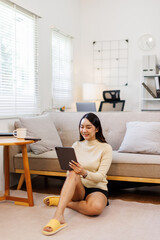 Attractive woman, freelancer holding and using digital tablet, sitting at cozy sofa in living room. Successful business asian woman text message in home. Online technology concept, remote job
