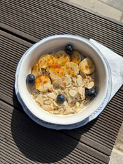 Oatmeal with Caramelized Bananas and Fresh Blueberries on Outdoor Table. Healthy Breakfast Out Concept. Handmade Mobile Photo, Harsh Sunlight, Not AI