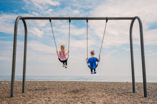 Kids swinging on a metal swing set at a pebbled beachside.