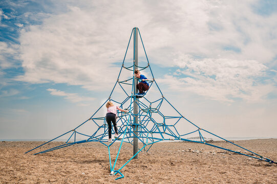 Two kids climbing a blue rope structure on a sandy beach.