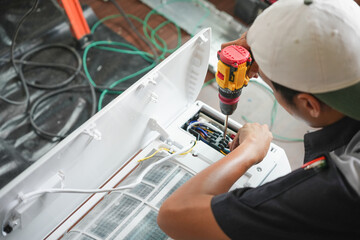 Technician Using Electric Drill to Repair Air Conditioning Unit, HVAC Technician Performing Maintenance on Indoor AC Unit, Young Worker Fixing Appliance with Cordless Drill Indoors