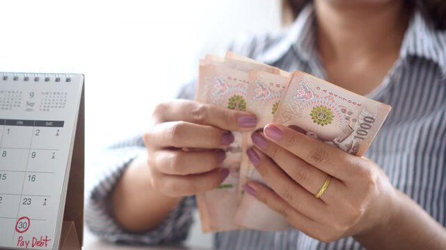 Close-up of a woman's hand counting Thai baht cash, with a reminder note on the calendar showing the due date for paying her debt or bills