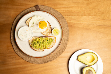 Top-down view of an avocado sandwich with toast and half-fried egg omelette on a white plate, alongside halved raw avocado on a small plate, set on a table in a restaurant.