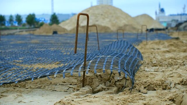 Installing plastic geogrid on sandy surface, reinforcing ground with metal rods and preparing terrain for upcoming asphalt paving process at infrastructure development site