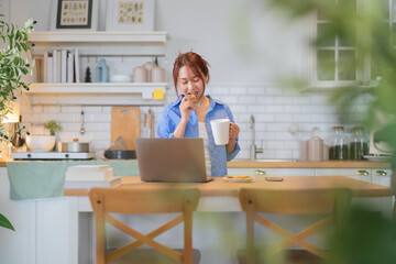 Young woman looking at laptop in kitchen  young Asian woman snacking on healthy rice crisps while working from home with laptop  working in kitchen with notebook 