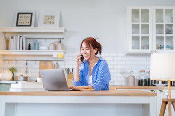 Obraz premium Asian adult Woman working from home in kitchen Beautiful young woman with an amputated arm working on a laptop and talking on a smartphone Woman Using Laptop At Home