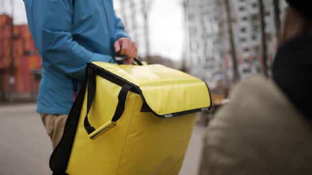 Food delivery courier wearing yellow thermal backpack handing over paper bag to customer outdoors, receiving cash payment during typical urban delivery transaction