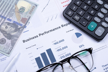 Business performance report with charts, calculator, pen, glasses, and US dollars on a desk, indicating financial analysis and planning.