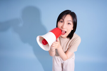 Young Asian woman enthusiastically using a megaphone against a blue backdrop in a creative studio setting, expressing excitement during a promotional activity