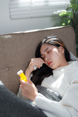 Sick young woman suffering from seasonal flu lying on sofa holding medicine