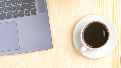 A laptop is open on a wooden table with a white coffee cup next to it. The coffee cup is filled with dark coffee, and the laptop is turned on. Concept of productivity and focus
