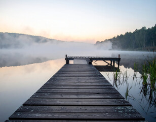 Fototapeta premium serene wooden pier extending into misty lake at dawn, surrounded by lush greenery and soft fog. tranquil atmosphere evokes sense of peace and calmness