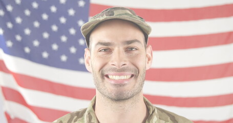 Smiling soldier in camouflage uniform and cap standing before American flag, with patriotic theme