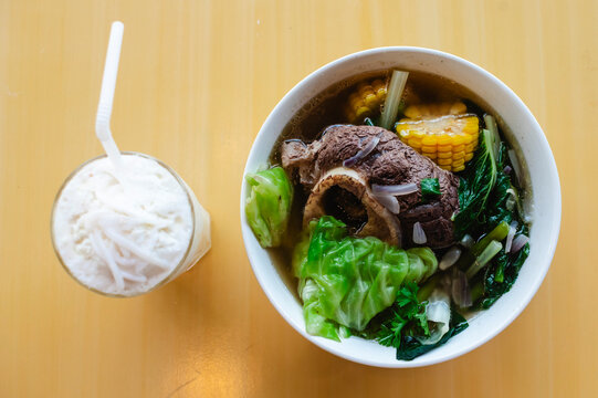 Top view of Filipino beef bulalo soup with leafy vegetables, corn, and bone marrow, served with chilled buko juice.