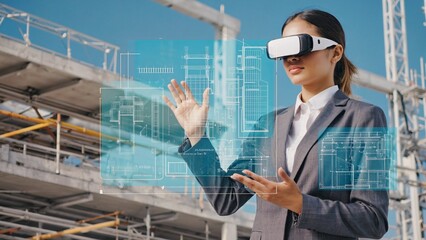 Asian female architect wearing a virtual reality headset is examining building blueprints using augmented reality technology at a construction site during a sunny day
