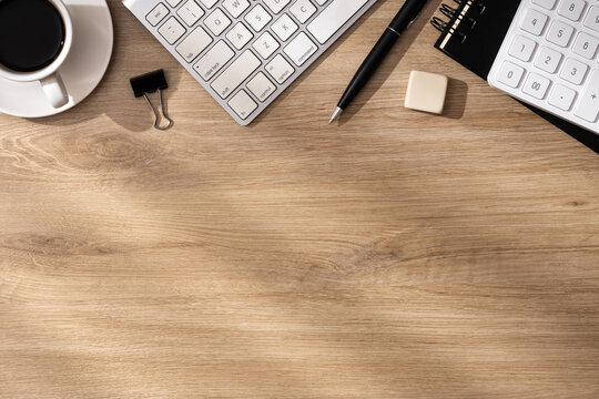 A morning workspace with a keyboard, notebook, coffee, calculator, pen and eraser on a wooden desk with sunlight from the window.