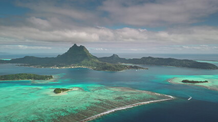 Naklejka premium Stunning aerial view of Bora Bora island with iconic mount Otemanu surrounded by turquoise water, coral reefs, and tropical greenery under cloudy sky. Remote wild nature paradise, exotic summer travel